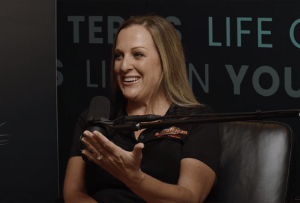 A woman sits in a podcast studio, smiling as she speaks into a microphone. She gestures with one hand while wearing a black polo shirt with a Bar-B-Clean logo. A dark background with partially visible text is behind her.