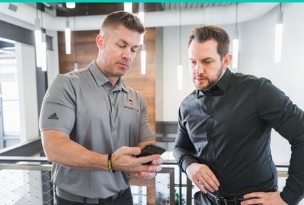 Two men stand in a modern office space, looking at a smartphone that one of them is holding. One man wears a gray polo shirt with a Sidekick logo, and the other wears a black button-down shirt. Bright hanging lights and industrial-style decor appear in the background.