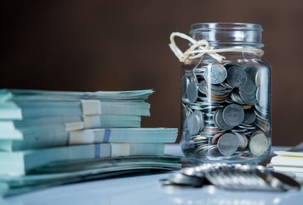 A glass jar filled with assorted coins sits on a reflective surface beside several neatly stacked bundles of paper money, with additional loose coins in the foreground.