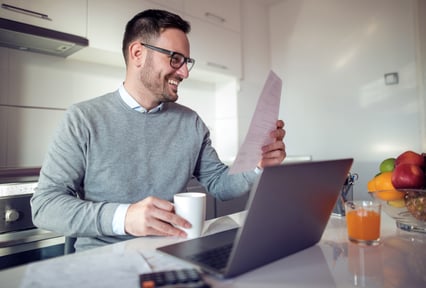 A guy sitting in front of a laptop holding a piece of paper and a mug, smiling