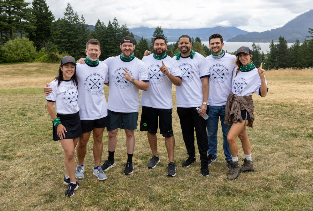 Attendees at a franchising event pose together outdoors in branded t-shirts with scenic mountain views behind them.