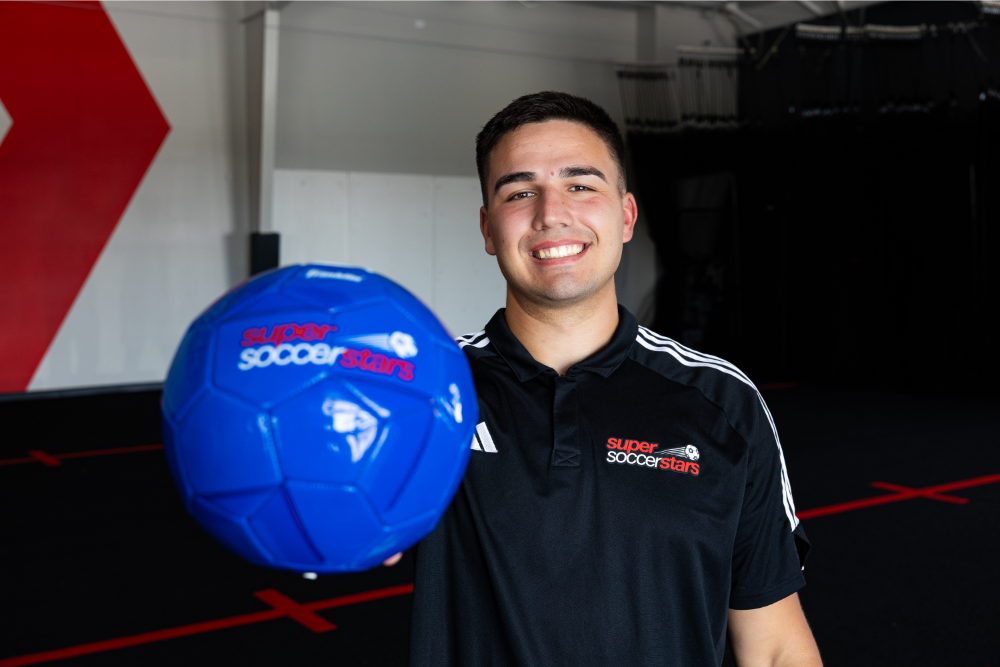 A smiling man wearing a black Super Soccer Stars shirt holds a bright blue soccer ball toward the camera inside an indoor sports facility. The background shows a large red graphic on the wall and a dimly lit training area.