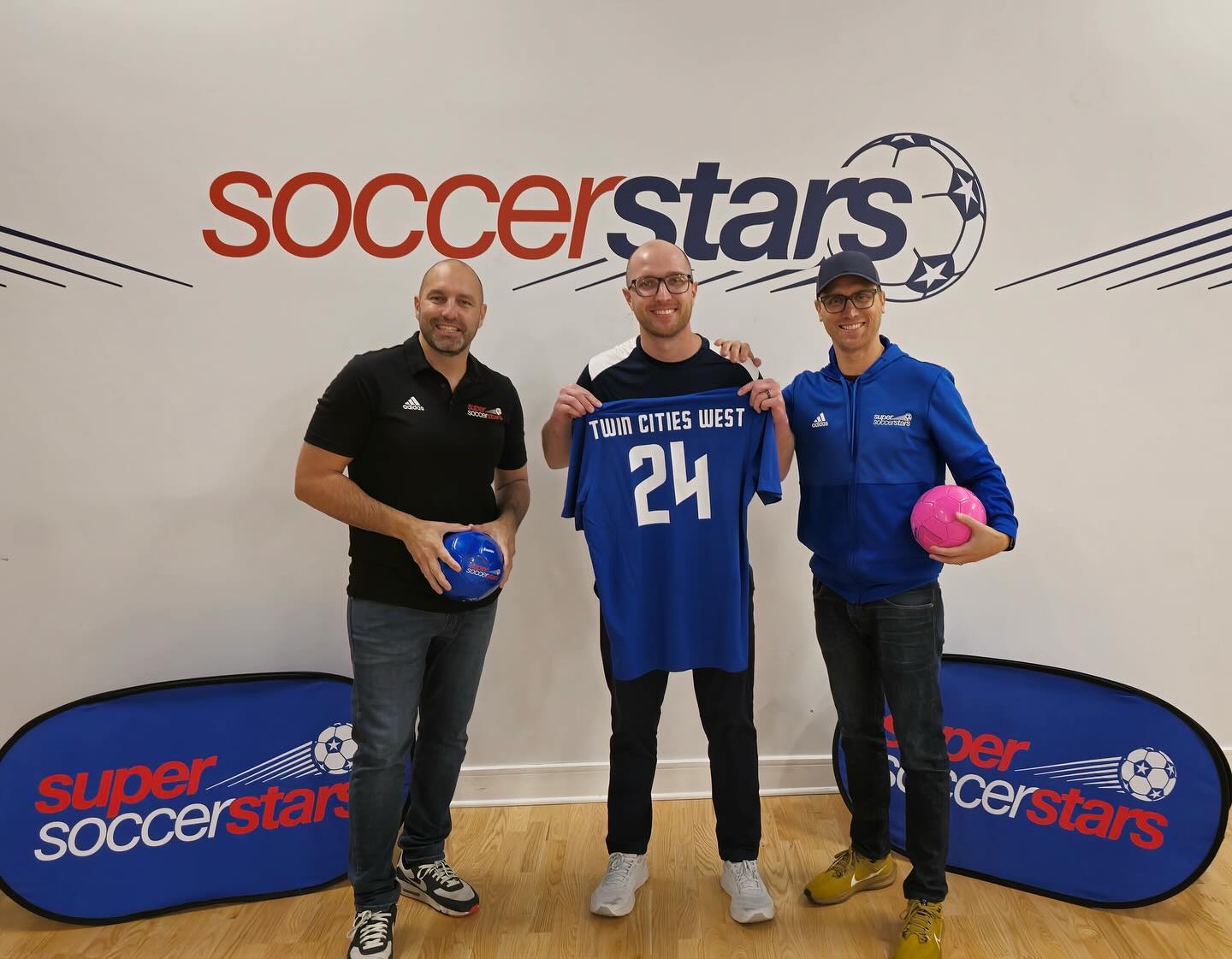 Three adults stand indoors in front of a wall with the “soccer stars” logo. The person in the center holds up a blue soccer jersey that reads “Twin Cities West” with the number 24. The two people on either side are smiling and holding soccer balls, with Super Soccer Stars banners displayed on the floor behind them.