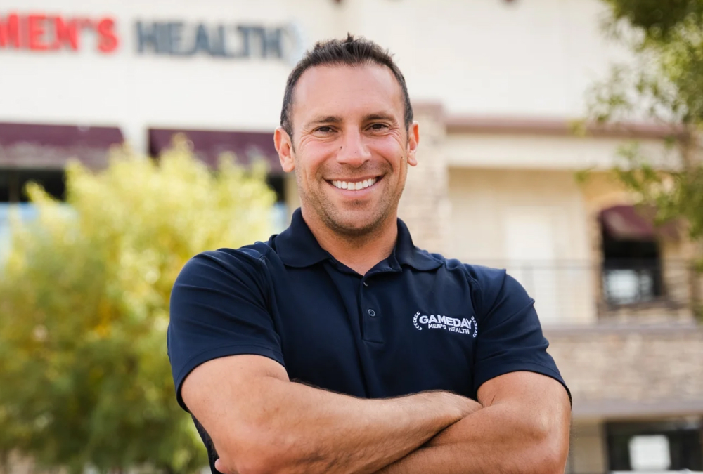 A smiling man in a navy GameDay Men’s Health polo stands outdoors with his arms crossed in front of a clinic building.