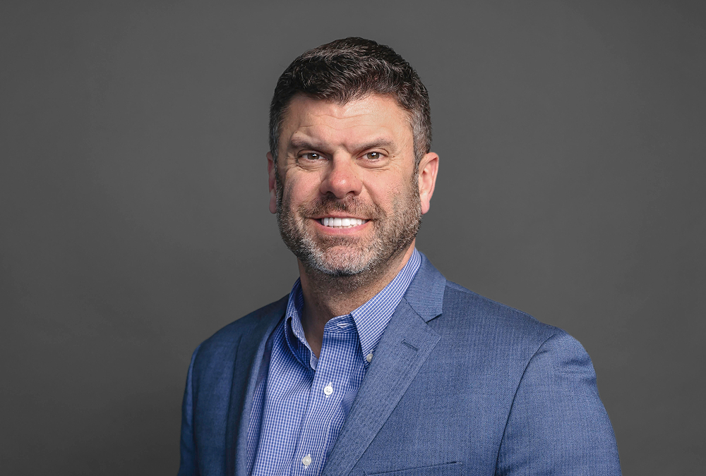 A man with short dark hair and a trimmed beard smiling at the camera. He is wearing a blue suit jacket over a light blue checkered shirt, posed against a plain gray background.