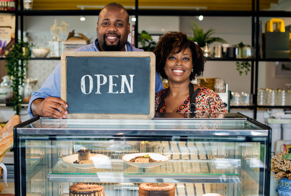 A smiling man and woman stand behind a bakery display case holding a chalkboard sign that reads “OPEN,” with cakes and pastries visible in the case and shelves of décor behind them.